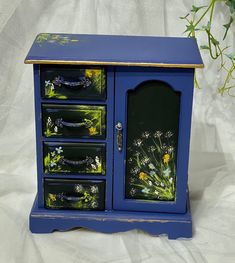 a small blue cabinet with flowers painted on it's doors and drawers, sitting next to a plant