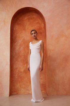 a woman standing in front of an orange wall wearing a white dress with one shoulder