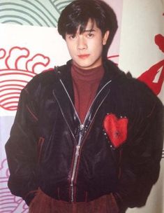 a young man standing in front of a wall wearing a black jacket and red shirt