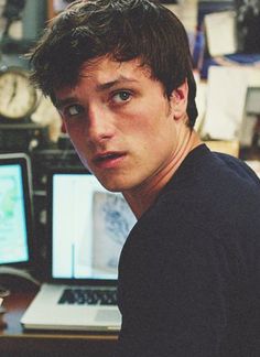 a young man sitting in front of a computer desk with two monitors and a clock on the wall