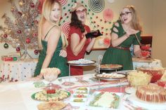 three women standing in front of a table full of food and desserts with christmas decorations on the wall behind them