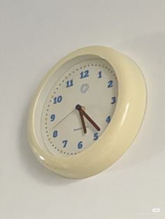 a white clock with blue numbers on the face and hands is hanging from a wall