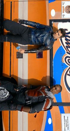 two men standing next to each other in front of a surfboard wall with an orange and blue background