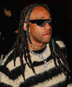 a man with dreadlocks wearing sunglasses and a striped sweater looks off to the side