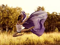 a woman is jumping in the air with a purple scarf on her head and dress blowing in the wind