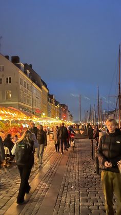 many people are walking on the sidewalk near some buildings and boats at night in the harbor