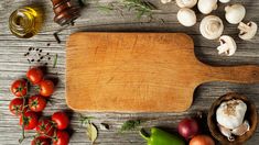 a cutting board surrounded by various vegetables and herbs on a wooden table with olives, tomatoes, garlic, peppers, mushrooms, and oil