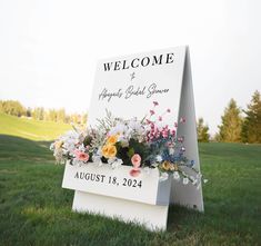 a welcome sign sitting in the grass with flowers on it's back and an open field behind it