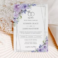 a wedding card with two rings on it and purple flowers in the middle, sitting next to some feathers