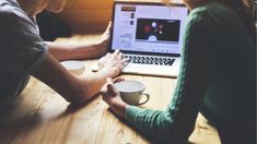 two people sitting in front of a laptop on a wooden table with coffee mugs