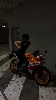 a woman sitting on an orange and white motorcycle in a parking garage with her hand on the seat