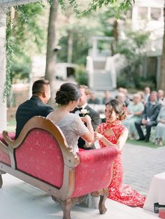 a bride and groom sitting on a bench in front of an outdoor wedding ceremony with guests