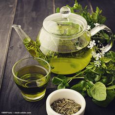 a glass tea pot filled with green liquid next to a cup of water and herbs