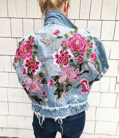 a woman wearing a jean jacket with pink flowers on the back and blue jeans, standing in front of a white brick wall