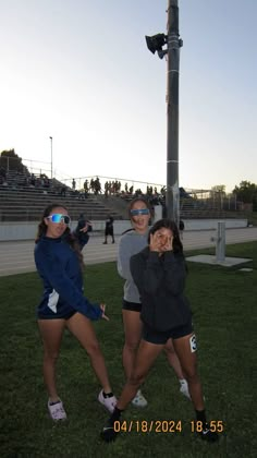 three girls are standing in the grass near a pole and street light with their hands up to their face