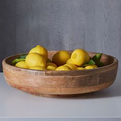 a wooden bowl filled with lemons on top of a white table next to a gray wall