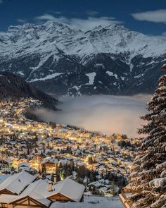 the town is surrounded by snow covered mountains and trees, with lights on in the foreground