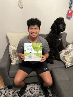 a boy sitting on a couch holding a book with a dog standing next to him