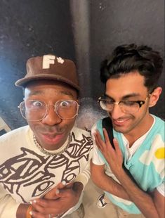 two young men posing for the camera with one holding his cell phone up to his face