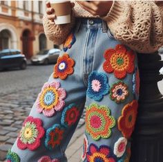 a woman standing next to a black pole wearing blue jeans with crocheted flowers on them