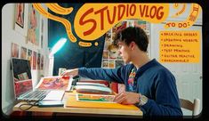 a young man sitting at a desk with a laptop computer in front of him and an advertisement for studio vlog on the wall behind him