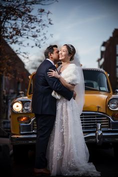 a bride and groom kissing in front of an old yellow car on the side of the road