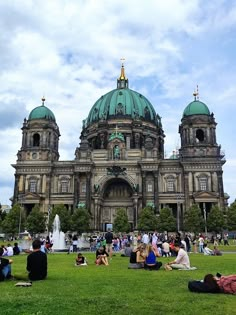 many people are sitting on the grass in front of a large building with green domes