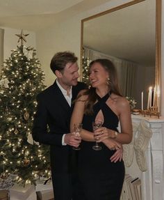 a man and woman standing in front of a christmas tree holding wine glasses smiling at each other