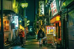 two people walking down an alley way at night with neon signs on the buildings in the background