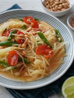 a white plate topped with noodles and veggies next to bowls of beans, limes and peanuts