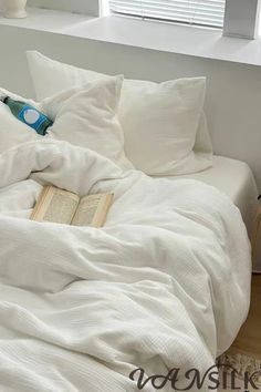 an unmade bed with white sheets and pillows in front of a window, reading a book