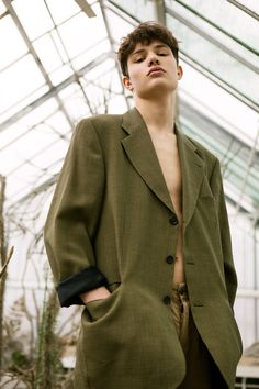 a man standing in front of a greenhouse wearing a green jacket and brown pants with his hands on his hips