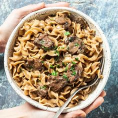 two hands holding a bowl of pasta with meat and mushrooms