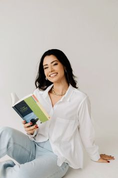 a woman sitting on the floor holding a book in her hands and smiling at the camera