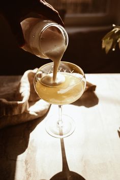 a person pouring something into a glass on top of a wooden table next to a potted plant