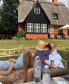 two people laying on a bench reading a book in front of a thatched roof house