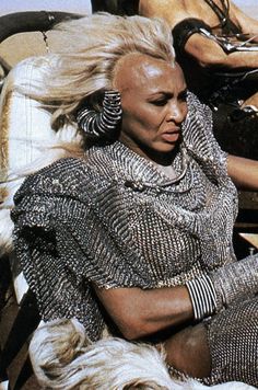 a woman sitting on top of a white horse next to two other women in black and silver outfits