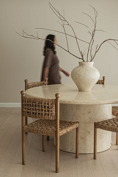 a woman is walking past a table with chairs and a vase on it in front of her