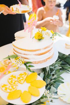 a white cake with yellow flowers on it and people in the background eating desserts