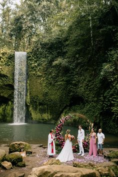 a group of people standing in front of a waterfall with flowers on the ground next to it