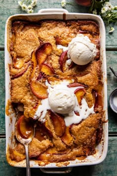 peach cobbler with ice cream on top in a white casserole dish next to flowers