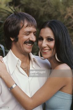 a man and woman hugging each other in front of some trees, smiling at the camera