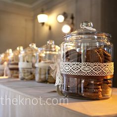 many jars filled with cookies on top of a white table cloth covered tablecloth and lights in the background