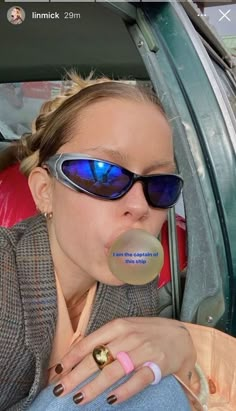 a woman wearing sunglasses and holding a bubble in her mouth while sitting in the back seat of a car