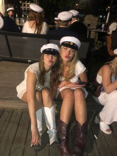 two women dressed in sailor hats sitting on a wooden deck with other people behind them