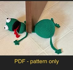 a crocheted green frog laying on the floor next to a door with its mouth open