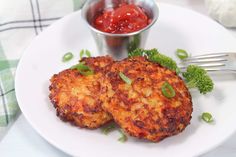 two crab cakes on a white plate next to a small bowl of ketchup