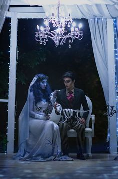 a man and woman dressed up in costumes sitting on a white chair under a chandelier