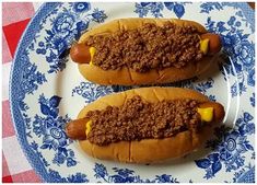 two hot dogs with chili on them sitting on a blue and white plate next to a checkered tablecloth