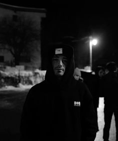 black and white photo of man in hoodie standing on sidewalk at night with other people